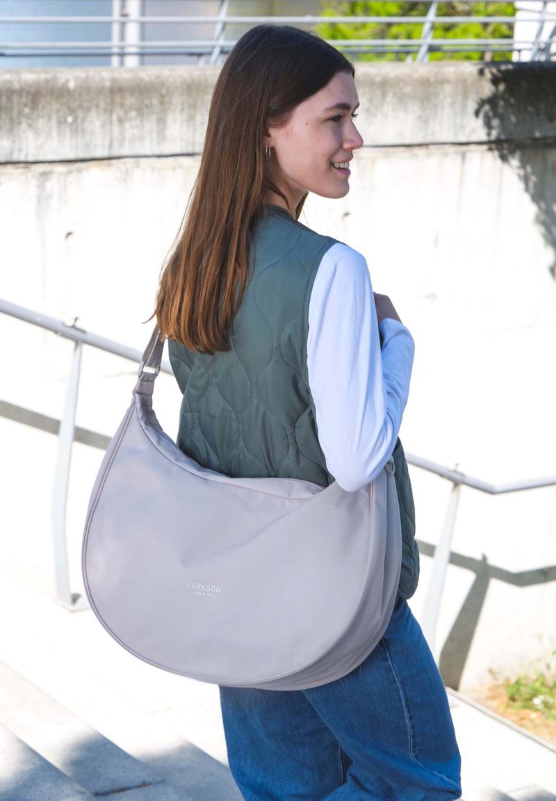 Borsa a spalla grigio chiaro realizzata in materiale morbido, con forma arrotondata e tracolla regolabile. Il modello indossa un gilet verde trapuntato e una camicia bianca.