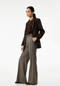 Bruine op maat gemaakte blazer over een donkere trui, gecombineerd met wijde, geruite broeken. Een bruine riem en subtiele oorbellen maken de look af.