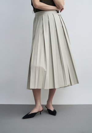 Femme portant une jupe midi plissée beige et des mules noires à talons bas et bouts pointus, debout contre un mur uni gris clair.