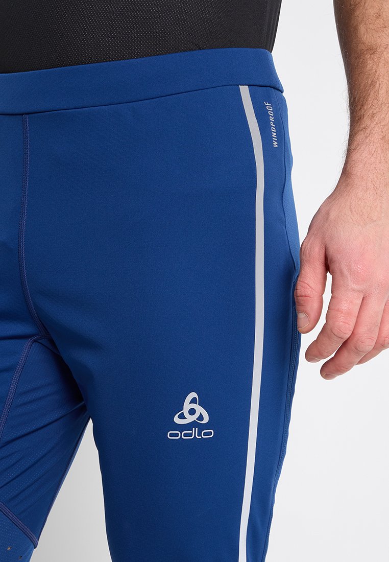 Zbliżenie na osobę w niebieskich sportowych spodniach od Odlo, odpornych na wiatr, z odblaskowymi paskami po bokach, oraz czarną górą.