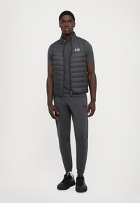 Gilet matelassé gris foncé sur une chemise à manches courtes assortie et un jogging. Design rembourré avec fermeture éclair avant et détail de logo. Baskets noires.
