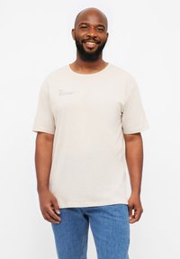 T-shirt en coton beige à manches courtes, col rond et design imprimé sur le devant. Présenté sur un fond blanc.