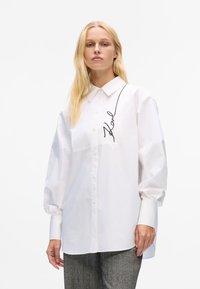White oversized shirt with button closure, structured collar, and black embroidered signature on the chest; smooth fabric and long sleeves.