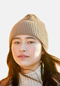 Braune gerippte Strickmütze mit spitzem Oberteil, getragen von einer Person. Weiche Textur; kombiniert mit einem cremefarbenen, hochgeschlossenen Pullover.