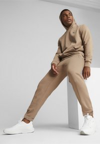 Felpa color sabbia con cappuccio e tasca frontale, abbinata a pantaloni coordinati, dotati di un logo decorativo. Scarpe da ginnastica bianche completano l'outfit.