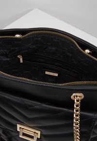 Czarna pikowana torebka na ramię ze złotym łańcuszkiem i zamkiem błyskawicznym, otwarta, pokazująca czarne wnętrze wyłożone wzorem z logo "ALDO" oraz wewnętrzną kieszeń na zamek.