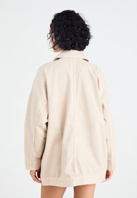 Beige oversized jakke i bomuldsstof, med en stor krave og droppe skuldre, med en glat tekstur og minimalistisk design.