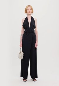 Svart halterneck jumpsuit med dyp V-hals, vid benform, glatt stoff, kombinert med en sølvfarget pailletveske og åpne svarte hæler.