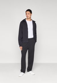 Μαύρο φούτερ με φερμουάρ και κουκούλα, φορεμένο πάνω από λευκό μπλουζάκι και μαύρη φόρμα. Το μοντέλο στέκεται με τα χέρια στις τσέπες, φορώντας λευκά αθλητικά παπούτσια.