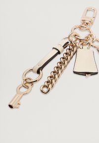 Porte-clés en ton doré avec une sangle en cuir, une chaîne maillon, une clé décorative et un onglet en cuir avec une texture lisse et des bords contrastés.