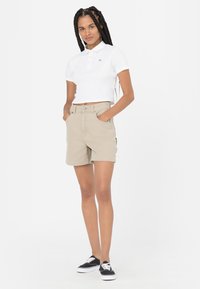 Maglietta polo bianca con un piccolo logo, abbinata a pantaloni corti beige dotati di tasche laterali. Modello che indossa Sneakers nere, in piedi contro uno sfondo bianco.