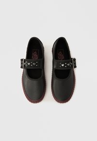 Fekete cipők sima szintetikus felsőrésszel, kerek orral és bordó texturált talppal. Ezüst szív díszítésekkel ellátott pánt jellemzi.