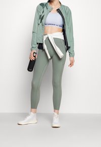 Ljusgrön atletisk jacka med svarta accenter, åtsittande ljusblå cropped topp, salviagröna leggings och vita träningsskor. Håller en svart vattenflaska.