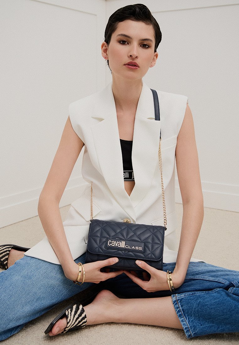 Bolso de mano acolchado en color navy con correa de cadena dorada y parche de marca. Presenta una forma rectangular y una textura suave con un ligero brillo.