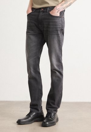 Man die versleten zwarte slim-fit jeans en zwarte leren nette schoenen draagt, met handen in de zakken en een zichtbaar getatoeëerde onderarm.
