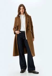 Manteau croisé en mélange de laine marron avec de grands boutons, porté sur une chemise blanche et un jean large bleu marine, associé à des mocassins noirs.