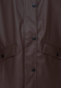 Imperméable marron vue de face en gros plan montrant des boutons-pression noirs et deux poches à rabat avec fermetures à pression de chaque côté.