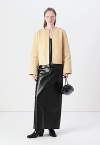 Veste bomber en cuir crème avec un col rond, associée à une longue jupe en cuir noir et des bottines noires à talons. Tenant un sac noir.