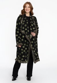 Zwarte omslag met groene cirkelvormige patronen, franjes aan de randen, lange mouwen en een open voorkant. Gekozen boven een zwarte outfit met strakke broek.