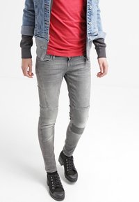 Los vaqueros skinny de denim gris presentan acentos de rodillas ribeteados y cinco bolsillos. Se llevan con una camiseta roja y una chaqueta de denim, combinados con zapatillas negras.
