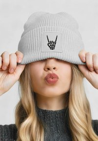 Gray ribbed knit beanie with a black skeleton hand graphic on a square patch, worn tilted down over a person's forehead.