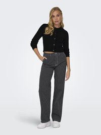 Schwarze gerippte Cropped-Cardigan mit Knöpfen, kombiniert mit hoch taillierten, gestreiften weiten Hosen. Das Model trägt weiße Sneaker und hat die Hände in den Taschen.