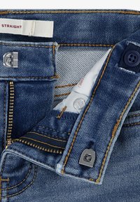 A farmernadrág egyenes szabású, kék színű, sárga varrással, fém cipzárral és deréknál gombos záródással rendelkezik.