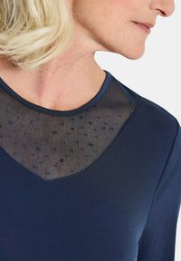 Top blu navy con scollatura a V in rete trasparente a pois. Tessuto morbido, design aderente e texture contrastanti. Cuciture visibili.