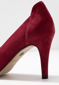 Červená semišová lodička s špičatou špičkou, úzkým stiletto podpatkem a hladkým vnitřním čalouněním. Obsahuje jemné prošívání a černou podešev.