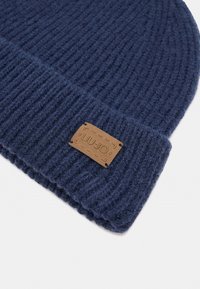 Beanie lavorato a maglia blu navy con una texture a coste e una patch in pelle beige con testo stampato in rilievo. Il design è semplice e accogliente.