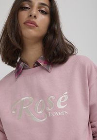 Felpa rosa con la scritta "Rosé Lovers" in testo argentato, sovrapposta a una camicia a quadri nei toni del rosa, bianco e viola. Texture del tessuto morbido visibile.