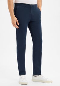 Pantalon en mélange de coton bleu marine avec une coupe slim, doté de poches latérales, d'une fermeture à bouton et d'une texture lisse. Associé à des baskets blanches.