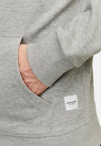 Grauer Hoodie mit einer Kängurutasche, gerippten Bündchen und einem Etikett mit "JACK & JONES EST. 1990", das an der unteren Vorderseite angenäht ist. Weiche Textur.