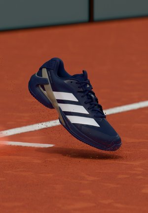 Scarpa da tennis Adidas blu navy con strisce bianche che solleva polvere di argilla su un campo da tennis in terra battuta rossa vicino a una linea di confine bianca.