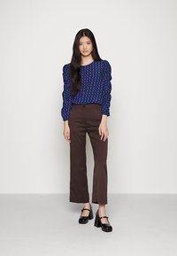 Blusa xadrez roxa e verde com mangas bufantes, combinada com calças retas castanhas e sapatos de salto bloco pretos.