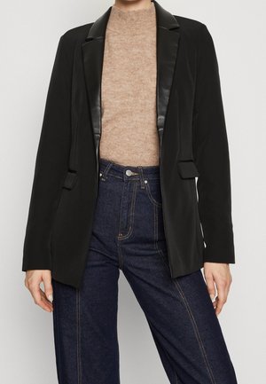 Blazer noir avec un revers en satin, manches longues, devant ouvert et poches latérales, porté sur un pull beige et un jean en denim bleu.