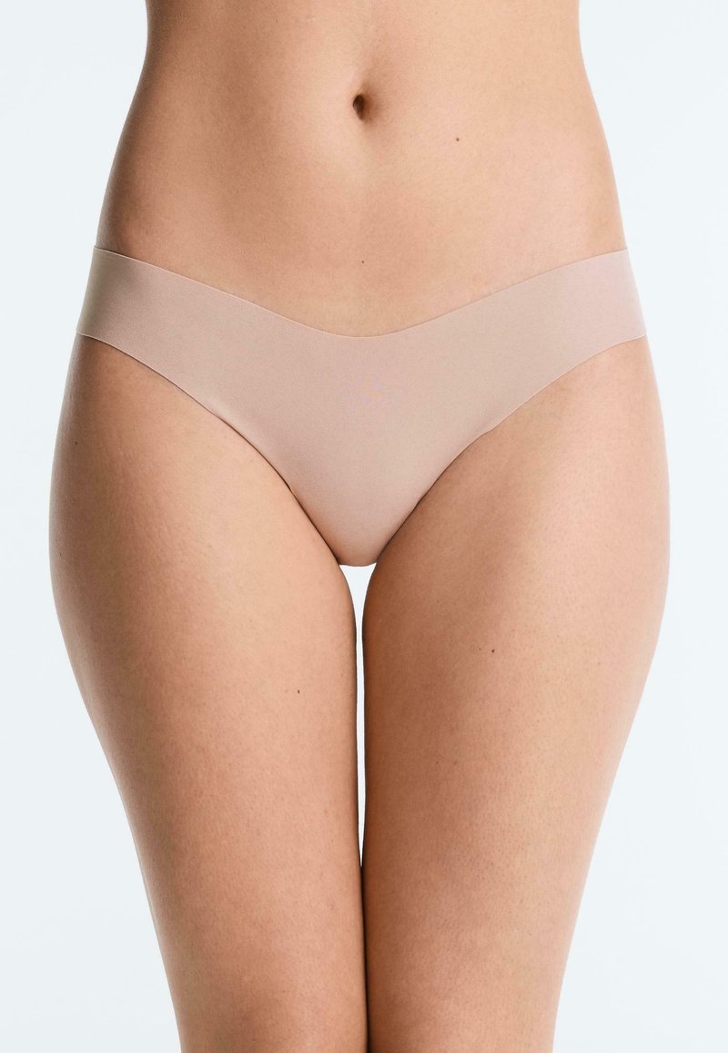 Bas du torse d'une femme portant une lingerie beige sans couture sur un fond clair uni.