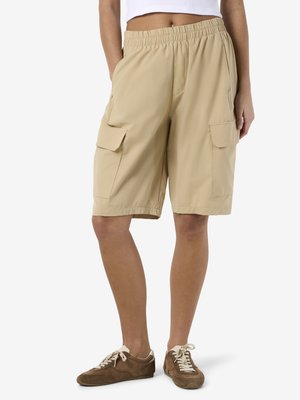 Osoba ubrana w beżowe szorty cargo do kolan z elastycznym pasem i kieszeniami, w połączeniu z brązowymi, casualowymi trampkami oraz krótką białą bluzką.