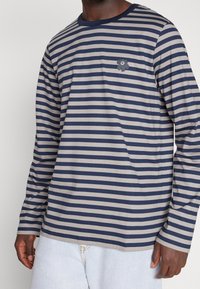 Randig långärmad skjorta i marinblå och grå, tillverkad av mjukt material. Har en liten blommönster på bröstet och rund halsringning.