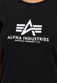 Juoda medvilninė marškinėliai su balta logo, kurioje yra raidė "A" ir trys horizontalių linijų virš jos, rodančios tekstą "ALPHA INDUSTRIES" ir vietos tekstą žemiau.