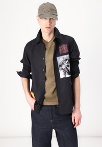 Black button-up shirt with three graphic patches, layered over an olive green t-shirt. Worn with dark jeans; cap with embroidered text.