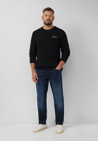 Homme debout contre un mur blanc portant un pull noir, un jean bleu et des baskets blanches, avec une main dans la poche.