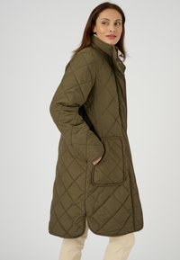 Abrigo largo acolchado color verde oliva con un alto cuello, dos bolsillos laterales y ribetes marrones en los bordes para un detalle y estructura adicionales.