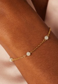 Goldene Kettenarmband mit drei abwechselnden weißen Perlen, auf einem braunen Hautton ruhend, vor einem sanften, neutralen Hintergrund.