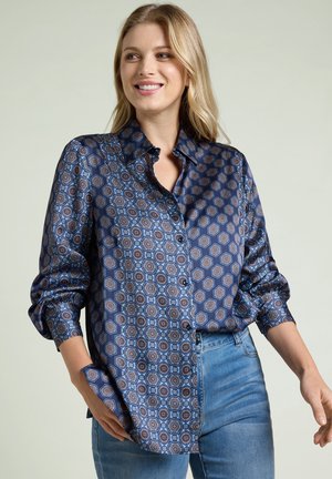 Leende kvinna klädd i en blå mönstrad knäppbar skjorta med geometriska mönster och ljusblå jeans, stående med ena handen i fickan.