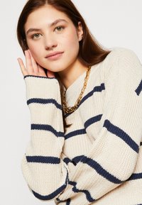 Pull en maille beige avec des rayures navy et des manches surdimensionnées. Il présente une texture côtelée et un col rond, assorti à un collier en chaîne dorée.