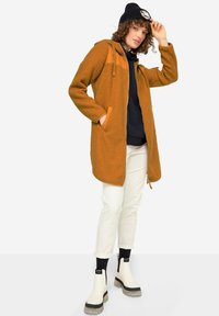 Fuzzy, bränd orange kappa med läderdetaljer, dragkedjestängning och huva, som bärs med vita byxor och svarta ankelstövlar.