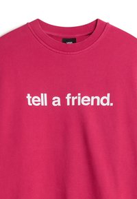Sweat-shirt rose vif à col rond avec texte blanc « tell a friend. » et une petite étiquette noire Vans au niveau du col.