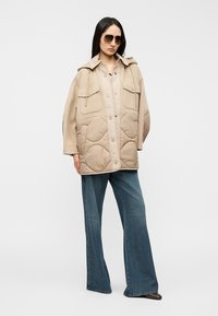 Bēša izšūta jaka ar kapuci, divām priekšējām kabatām, spiedpogām un ērtu siluetu. Kombinēta ar platām zilām džinsa biksēm.