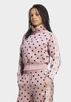 Femme aux cheveux tressés portant un survêtement Adidas rose clair à pois avec des bandes blanches, les mains dans les poches, regardant devant avec confiance.